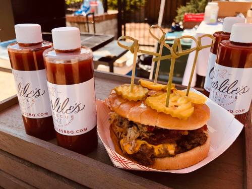 A hearty burger topped with pickles and cheese, accompanied by bottles of barbecue sauce in the background.