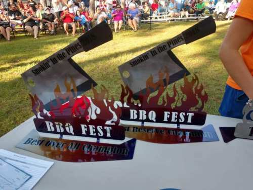 Two colorful BBQ fest trophies with flames, displayed on a table at an outdoor event, surrounded by an audience.