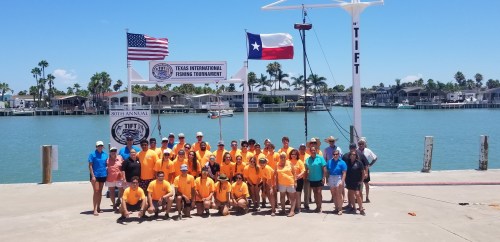 Group photo of participants at the Texas International Fishing Tournament by the water, with flags and a banner in the background.
