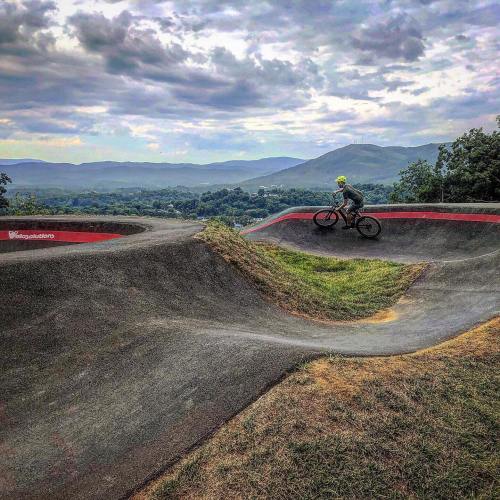 A cyclist rides on a dirt pump track surrounded by green hills and cloudy skies.