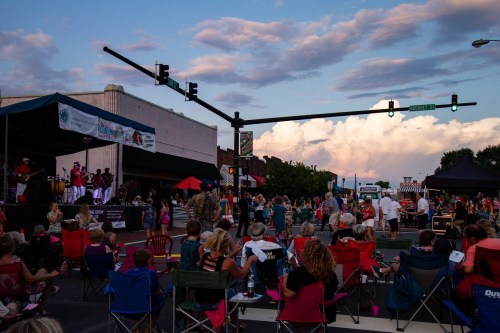 A lively street festival with crowds sitting in chairs, a stage with performers, and colorful tents under a cloudy sky.