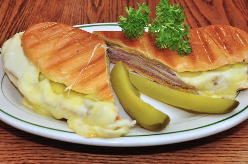 A toasted sandwich filled with melted cheese and ham, served with pickles and garnished with parsley.