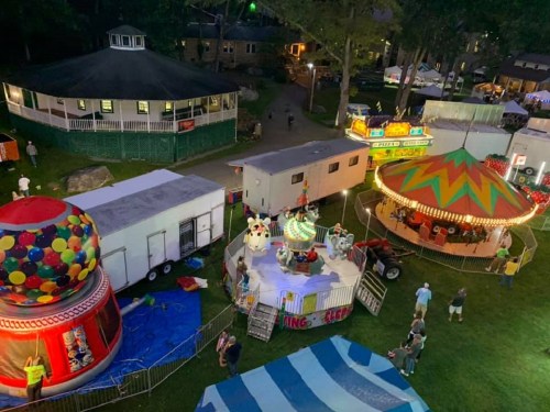 Aerial view of a carnival with colorful rides, a carousel, and food trailers surrounded by trees at night.