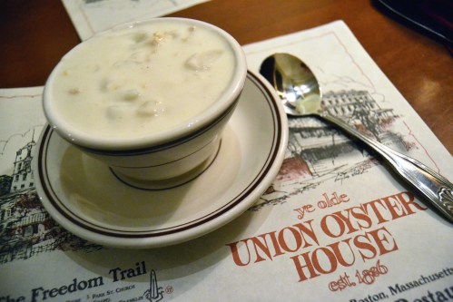 A bowl of clam chowder on a plate, next to a spoon, with a menu from Union Oyster House in the background.