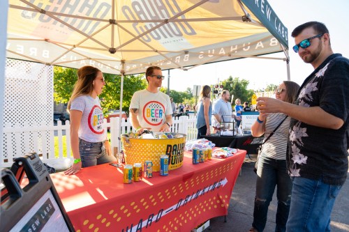 Two people at a booth with a sign reading "Beer Me," serving drinks to a customer at an outdoor event.