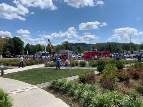 A busy outdoor area with food trucks, people enjoying the day, and green landscaping under a blue sky with clouds.