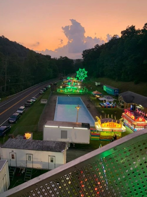 Aerial view of a carnival at sunset, featuring colorful lights, rides, and a pool surrounded by trees and parked cars.