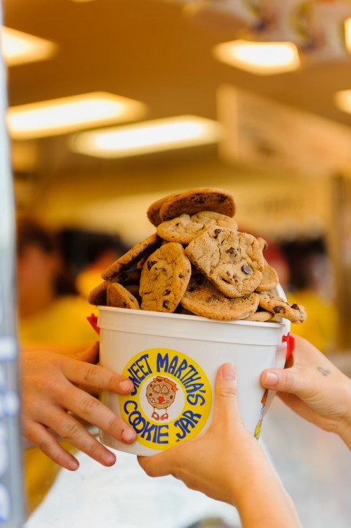 A bucket overflowing with chocolate chip cookies, with hands reaching in to grab some.