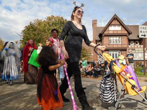 A woman in a black outfit walks in a parade, holding a child's hand, with costumed participants and a stroller nearby.