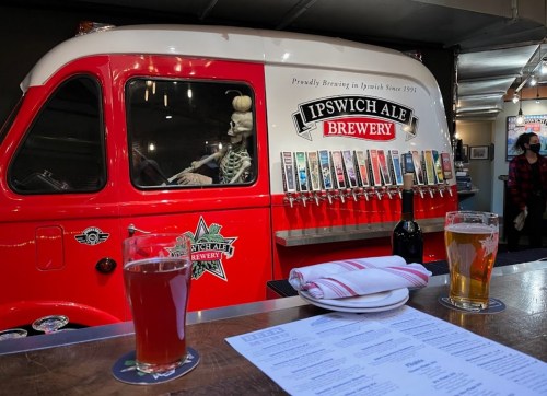 A vibrant brewery bar with a skeleton in a red truck, showcasing various beer taps and two glasses of beer in the foreground.