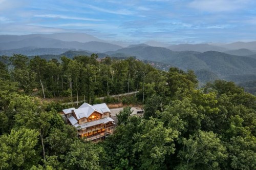 Aerial view of a wooden cabin nestled among lush green trees, surrounded by misty mountains under a blue sky.