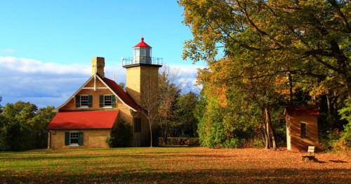 A scenic lighthouse beside a charming brick house, surrounded by colorful autumn foliage and a clear blue sky.
