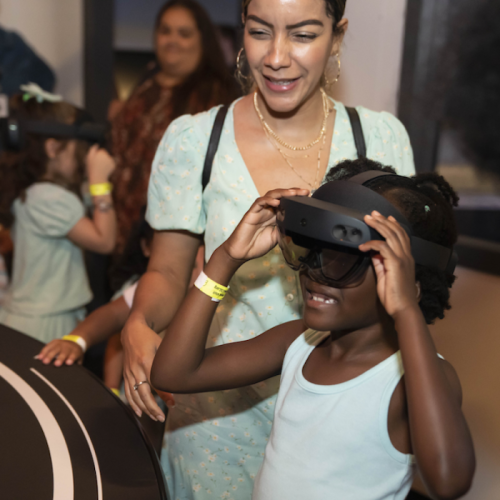 A woman assists a young girl wearing a virtual reality headset at an event, with other children in the background.