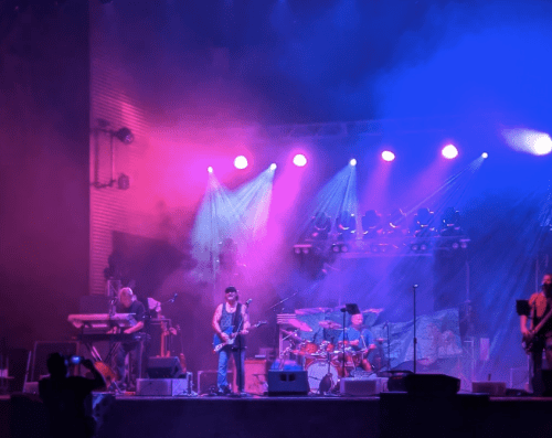 A vibrant live music performance with a guitarist, drummer, and keyboardist under colorful stage lights and smoke.