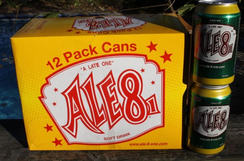 A yellow box of 12-pack Ale-8-1 soft drink cans next to two individual cans, with a blue background.