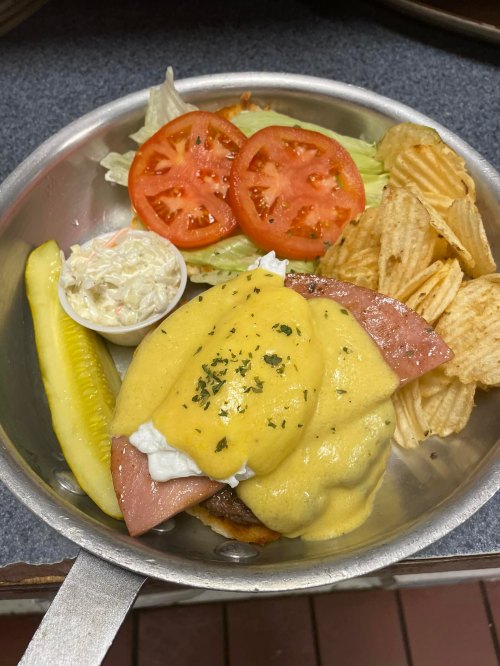 A plate with a sandwich topped with hollandaise sauce, pickles, lettuce, tomato, coleslaw, and potato chips.
