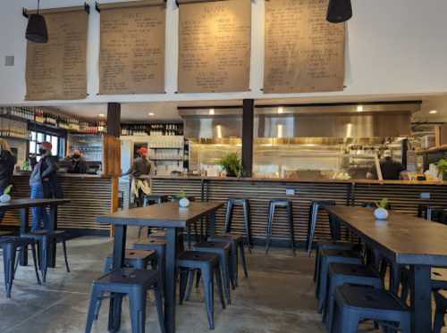 Interior of a restaurant featuring wooden tables, metal chairs, a menu board, and an open kitchen area.