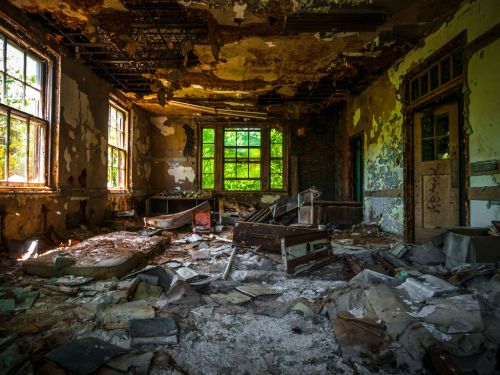 Abandoned room with peeling paint, broken furniture, and debris scattered across the floor, illuminated by natural light.