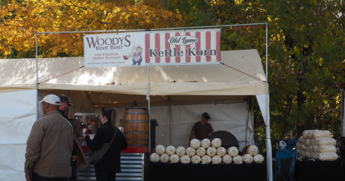 A vendor tent selling Woody's Root Beer and Old Tyme Kettle Korn, with customers browsing in a fall setting.