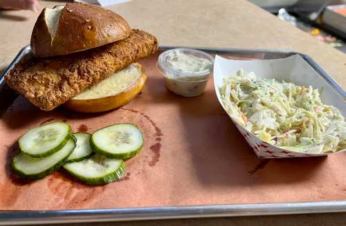 A plate featuring a fried fish sandwich on a pretzel bun, pickles, coleslaw, and a small cup of sauce.
