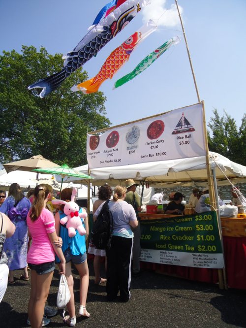 A food stall at a festival with colorful banners, offering various dishes and snacks, and a line of customers.