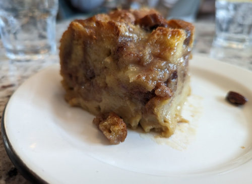 A slice of bread pudding on a white plate, with a rich, creamy texture and bits of chocolate visible.