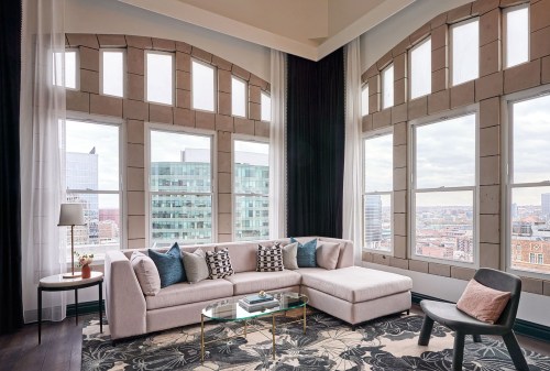 Bright, modern living room with large windows, a light sofa, decorative pillows, and a glass coffee table overlooking the city.