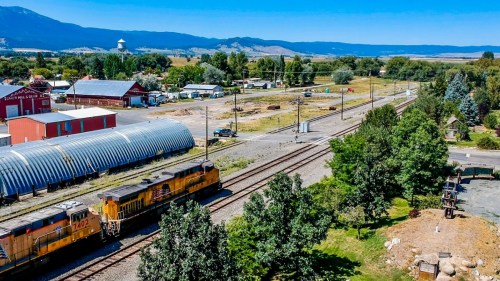 Aerial view of a rural area with train tracks, a yellow locomotive, and buildings surrounded by trees and mountains.