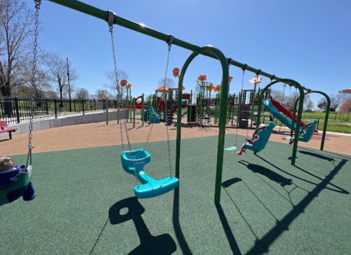 A sunny playground featuring swings, slides, and climbing structures on a rubberized surface.