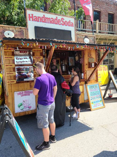 A wooden food stall labeled "Handmade Soda" with colorful signs, people ordering drinks on a sunny day.