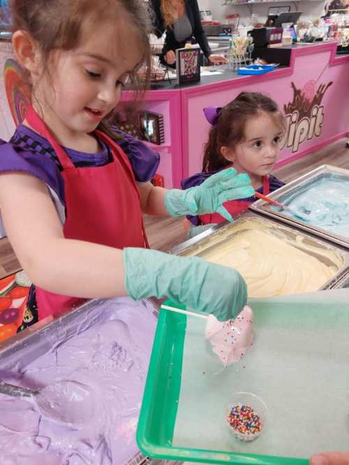 Two young girls in aprons are decorating ice cream with colorful sprinkles at a vibrant ice cream shop.