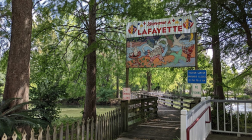A colorful sign reading "Bienvenue à Lafayette" above a wooden walkway leading to a green park area.