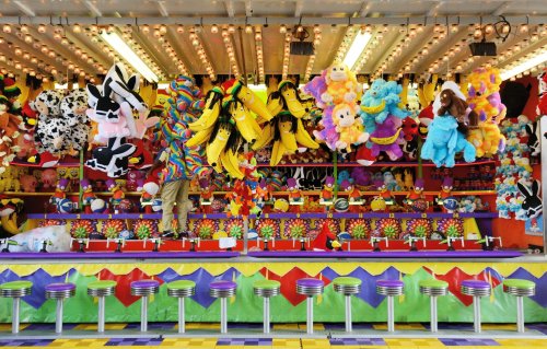 A colorful carnival game booth filled with plush toys and prizes, with bright lights and empty stools in front.
