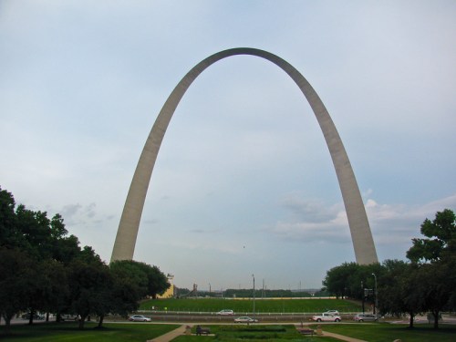 A tall, curved monument stands against a cloudy sky, surrounded by green grass and trees. Cars are visible in the foreground.
