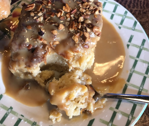 A slice of cake topped with granola and drizzled with a creamy sauce, served on a green-checkered plate.
