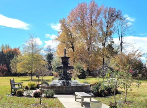 A serene park scene featuring a fountain surrounded by trees with autumn foliage and benches on a sunny day.