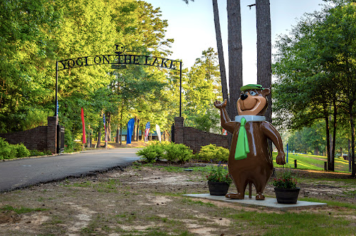 A bear statue in a green hat waves at the entrance of a park, surrounded by trees and colorful flags.