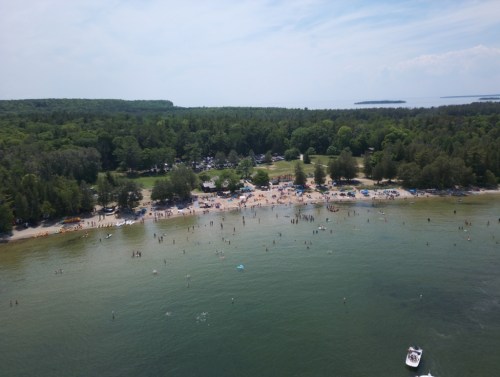 Aerial view of a busy beach with people swimming, sunbathing, and trees lining the shore under a clear sky.