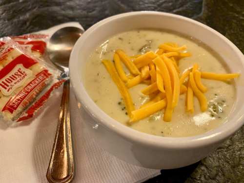 A bowl of creamy soup topped with shredded cheese, accompanied by a packet of saltine crackers and a spoon.