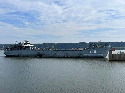 A large gray military ship docked in calm waters, with hills and a cloudy sky in the background.