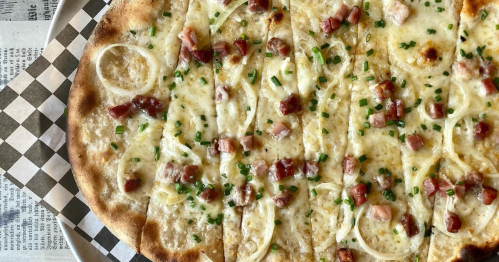 A close-up of a pizza topped with cheese, diced ham, onions, and green chives on a checkered paper background.