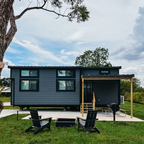 A modern gray tiny house with a porch, surrounded by green grass and trees, under a cloudy sky.
