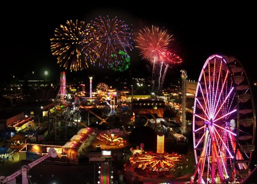 Colorful fireworks illuminate a vibrant carnival at night, featuring a Ferris wheel and various rides.