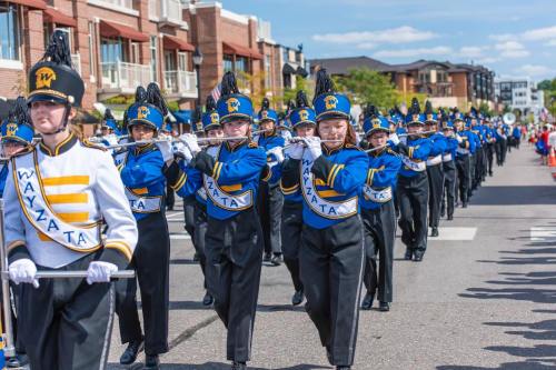 A marching band in blue and white uniforms parades down a street, with musicians playing instruments and smiling.