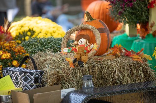 A festive autumn display featuring a scarecrow, pumpkins, and colorful flowers on a hay bale.