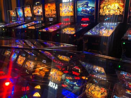 A vibrant arcade filled with pinball machines, reflecting colorful lights and artwork on their surfaces.