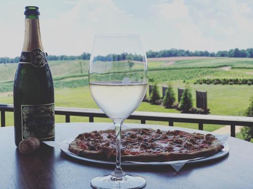 A glass of white wine beside a pizza on a table, with a vineyard landscape in the background.