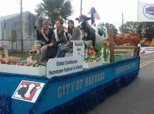 Three women in sashes wave from a decorated float at a parade, with a cow statue and autumn decorations.