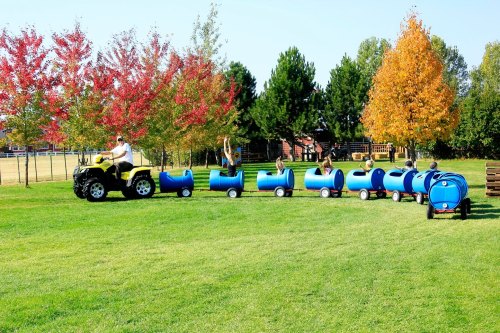 A person drives an ATV pulling a colorful train of barrels, with children enjoying a ride on a sunny day in a park.