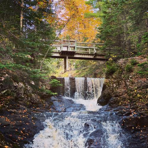 A wooden bridge spans a waterfall surrounded by autumn foliage and rocky terrain.